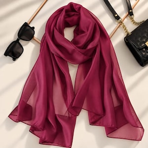 Puede incluir: Una bufanda de color magenta sólido hecha de una tela ligera y semitransparente con una textura suave. La bufanda está drapeada en un bucle central, mostrando su color vibrante y el material fluido y translúcido que crea pliegues suaves.