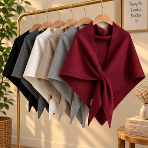 May include: A collection of six triangular knit shawls hanging on a gold metal rack. The shawls are displayed in a gradient of colors including black, dark gray, white, beige, light gray, and a deep burgundy red, each featuring a small side tag.