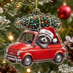 May include: A clear acrylic Christmas ornament shaped like a red vintage car with a raccoon wearing a Santa hat labeled Oliver driving. The car carries a decorated Christmas tree on its roof, featuring colorful ornaments and a gold star topper.