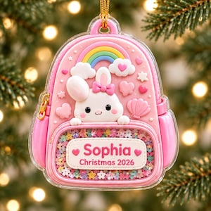 May include: A pink backpack-shaped Christmas ornament featuring a white bunny with a pink bow, a rainbow, clouds, and hearts. The front pocket is a clear shaker filled with colorful flower sequins and a label reading Sophia Christmas 2026.