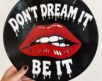 May include: A black vinyl record featuring a graphic of bright red lips with white teeth. The white text above the lips reads "DON'T DREAM IT" and the white text below the lips reads "BE IT". The lettering has a dripping paint effect.