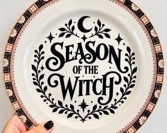 May include: A round ceramic plate with a cream-coloured centre featuring the black text Season of the Witch, surrounded by a black crescent moon, stars, and leafy vines. The rim has a decorative border of black, white, and orange geometric patterns.