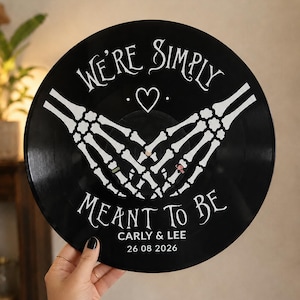 May include: A black vinyl record featuring white printed graphics of two skeleton hands forming a heart shape. The text reads WE'RE SIMPLY MEANT TO BE, with the names CARLY & LEE and the date 26 08 2026 printed in white below the skeleton hands.