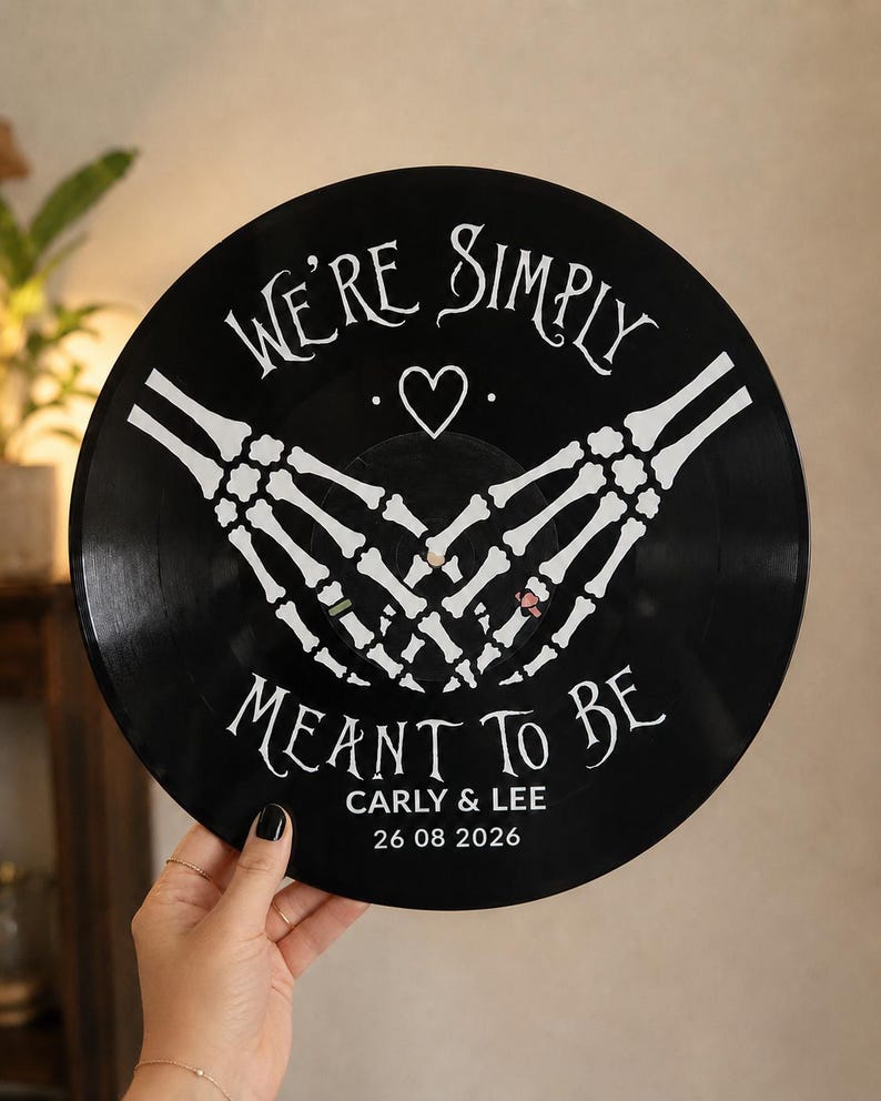 May include: A black vinyl record featuring white printed graphics of two skeleton hands forming a heart shape. The text reads WE'RE SIMPLY MEANT TO BE, with the names CARLY & LEE and the date 26 08 2026 printed in white below the skeleton hands.