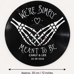 May include: A black vinyl record wall clock featuring white skeleton hands forming a heart shape. The text reads WE'RE SIMPLY MEANT TO BE, CARLY & LEE, and 26 08 2026. A double-headed arrow indicates a diameter of approximately 30 cm.