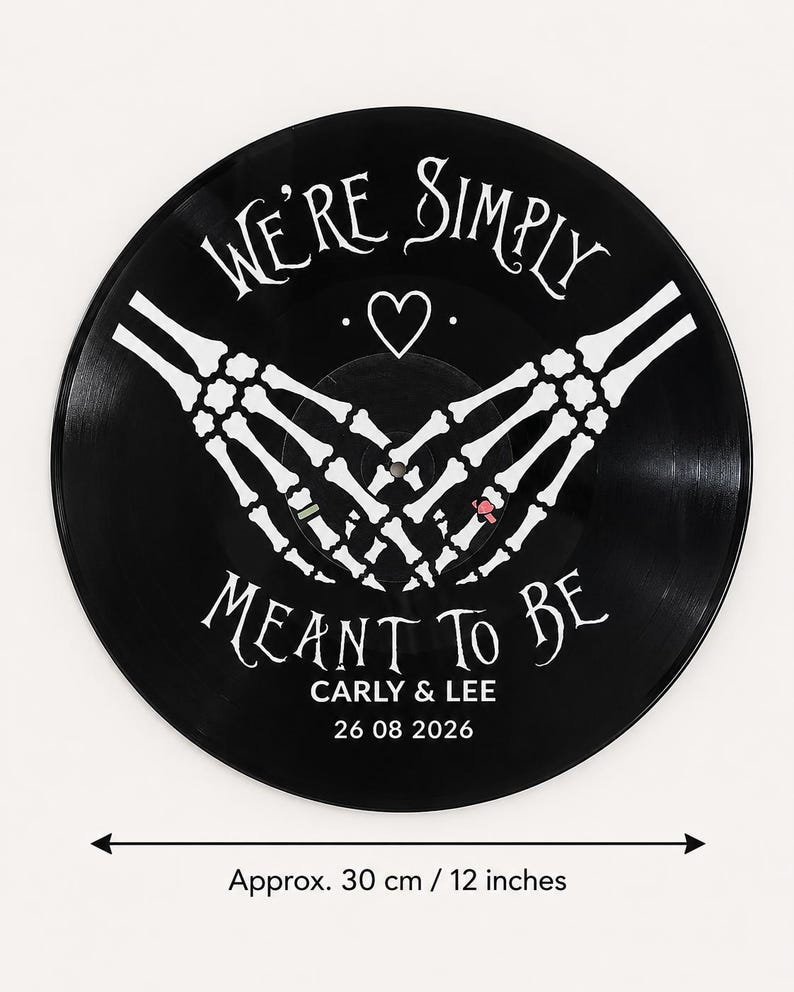 May include: A black vinyl record wall clock featuring white skeleton hands forming a heart shape. The text reads WE'RE SIMPLY MEANT TO BE, CARLY & LEE, and 26 08 2026. A double-headed arrow indicates a diameter of approximately 30 cm.