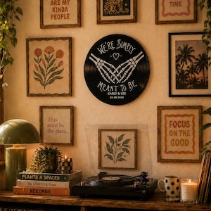 May include: A circular black vinyl record wall art piece featuring white skeletal hands forming a heart shape with the text "WE'RE SIMPLY MEANT TO BE" above and "CARLY & LEE 26 08 2026" printed below, displayed on a wall surrounded by framed prints.