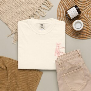 May include: A cream-colored short-sleeve t-shirt with a crew neckline. The shirt features a small graphic on the lower right side depicting a pair of light pink ballet slippers with ribbons and the text "Dance Mom" in a cursive script font.