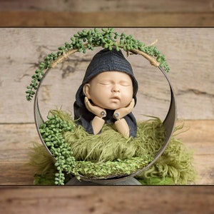May include: A sleeping baby doll wearing a blue knitted hat and a green fuzzy blanket, posed inside a metal hoop decorated with green leafy vines.