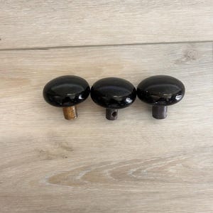 May include: Three round, glossy black knobs with smooth, domed surfaces. Each knob features a short, cylindrical base extending downward, with the center knob showing a small circular hole in its metallic mounting stem for attachment purposes.