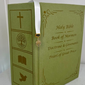 May include: A green hardcover book titled Holy Bible, Book of Mormon, Doctrine & Covenants, and Pearl of Great Price. The spine features gold icons of a cross, tree, open book, and dove. A white ribbon bookmark hangs with a gold tree charm.