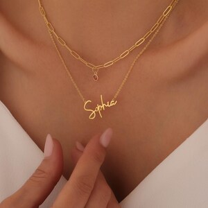 May include: A gold-toned layered necklace set featuring a top chain with elongated oval links and a small round red stone charm, and a bottom chain with a cursive nameplate pendant displaying the name Sophia in a script font style.