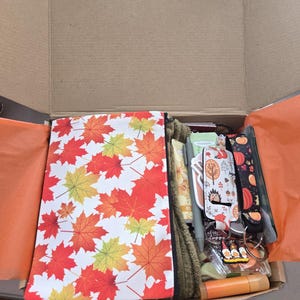 May include: A cardboard gift box containing a zippered pouch with a red, orange, and yellow maple leaf pattern, a green textured fabric, a gray pen, a lip balm, a keychain with a pumpkin print, and a small charm featuring three gnomes.