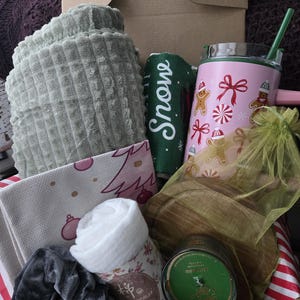 May include: A gift box containing a sage green waffle-knit blanket, a pink tumbler with gingerbread and candy cane prints, a green towel with the word Snow, a white cloth, a grey scrunchie, a green candle, and a small yellow organza bag.