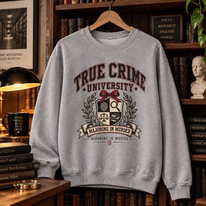 Può includere: Una felpa girocollo grigio melange con una stampa grafica che recita True Crime University, Majoring in Murder e Minoring in Motive. Il design include uno scudo con un'impronta digitale, una lente d'ingrandimento, una bilancia e libri.