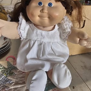 May include: A doll with brown hair and blue eyes wearing a white dress with ruffled sleeves and white fabric shoes. The doll has a round face with rosy cheeks and a soft, fabric-based body. The dress features a simple round neckline.