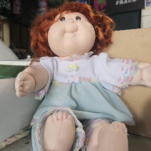 May include: A doll with auburn hair and a round face wearing a white top with short sleeves and a light blue skirt. The outfit features multicoloured lace trim at the neckline, sleeves, and hem. The doll has a soft fabric body and light skin tone.