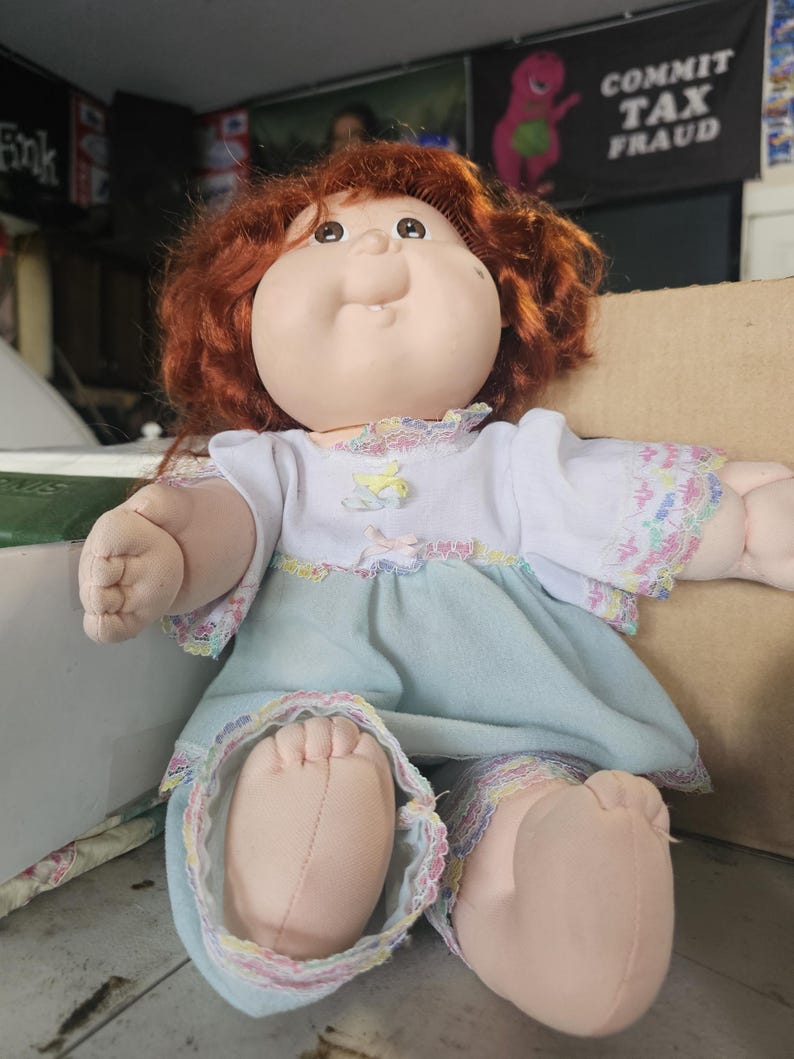 May include: A doll with auburn hair and a round face wearing a white top with short sleeves and a light blue skirt. The outfit features multicoloured lace trim at the neckline, sleeves, and hem. The doll has a soft fabric body and light skin tone.