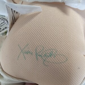 Op de afbeelding: Een beige stoffen pop met een textuur op het oppervlak, voorzien van een groene handtekening van Xavier Roberts aan de onderkant. De pop draagt een wit kledingstuk met een subtiel stippenpatroon en heeft een wit stoffen label met tekst.