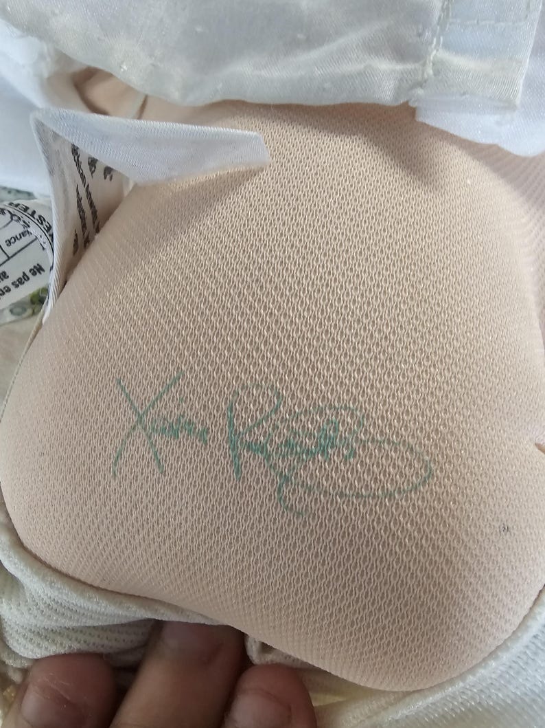 Op de afbeelding: Een beige stoffen pop met een textuur op het oppervlak, voorzien van een groene handtekening van Xavier Roberts aan de onderkant. De pop draagt een wit kledingstuk met een subtiel stippenpatroon en heeft een wit stoffen label met tekst.