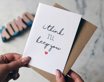 May include: A white card with a handwritten message that says "i think i'll keep you" with a small red heart below the text. The card is held in a person's hands.