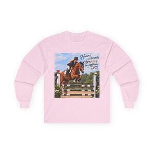 May include: A light pink long-sleeve shirt featuring a graphic print of a rider on a brown horse jumping over a hurdle. The text "Hooves in the air, dreams in motion" is printed in dark blue script next to the horse, along with a small horse icon.