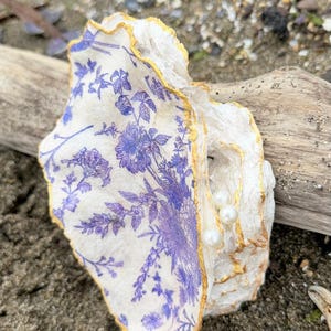 May include: A decorative oyster shell featuring a blue floral pattern on its interior surface, accented with gold-painted edges. Three small, round, white pearls are attached to the inner shell, which rests against a piece of weathered driftwood.