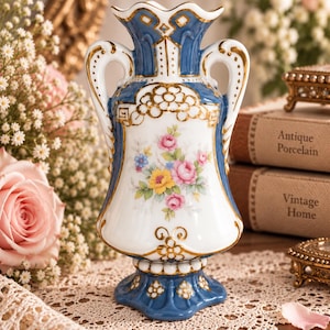 Può includere: Un vaso in porcellana con corpo bianco e un motivo floreale dipinto in blu e oro. Il vaso presenta due manici, un bordo smerlato e una base svasata. Il design centrale mostra fiori rosa, gialli e blu con foglie verdi e dettagli dorati.