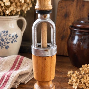 Op de afbeelding: Een vintage houten pepermolen met een lichtbruine houten voet en een bijpassende houten knop op de hendel. Het centrale mechanisme is gemaakt van verweerd zilverkleurig metaal dat de houten hendel verbindt met het hoofdgedeelte van de molen.
