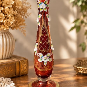 May include: A tall, slender red glass vase featuring a gold-rimmed opening and base. The body is decorated with intricate gold filigree, a lattice pattern, and raised, hand-painted white and blue floral motifs with green leaves on the exterior.