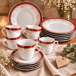 Op de afbeelding: Een set witte porseleinen theekopjes en schoteltjes met een dieprode rand en ingewikkelde gouden filigraanpatronen. De collectie bevat gestapelde borden en kopjes, met een consistent decoratief motief van sierlijke gouden krullen op rood.