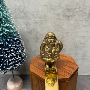 May include: A brass-colored Santa Claus figurine with a long beard and a sack over his shoulder. The figurine is attached to a gold-colored bar. A small, blue Christmas tree is in the background.