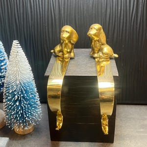 May include: Two brass dachshund dog stocking holders. Each holder features a seated dog figure atop a curved, gold-colored metal piece. The holders are displayed on a dark wooden block, with a dark background.