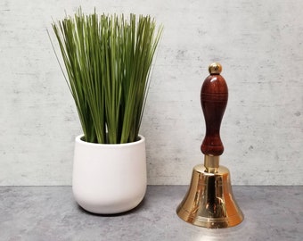 May include: A brass bell with a wooden handle. The bell is gold and has a rounded shape. The handle is dark brown and has a smooth finish. The bell is sitting on a gray surface next to a white planter with green grass.