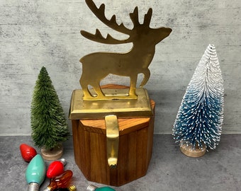 May include: A gold reindeer stocking holder with a wooden base. The holder is decorated with a small, green pine tree and a blue and white pine tree. There are also several colorful light bulbs scattered around the base.
