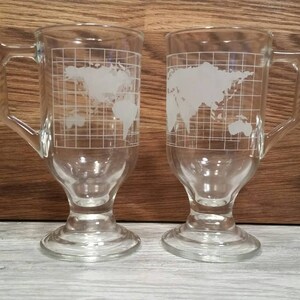 May include: Two clear glass mugs with handles, each featuring a white etched world map design. The mugs have a footed base.