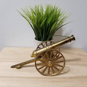 May include: A brass cannon model with spoked wheels and a long barrel. The miniature cannon is displayed on a wooden surface, with a green plant in a white pot in the background. The cannon is a decorative item.
