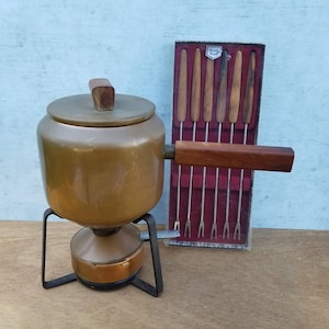 May include: A vintage copper fondue pot with a wooden handle and a stand. The pot has a lid and is on a black metal stand. The pot is accompanied by a box of six fondue forks with wooden handles. The box has the text "Hostess" printed on it.