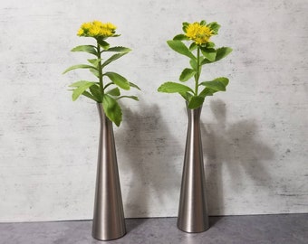 May include: Two silver vases with a yellow flower arrangement in each. The vases are tall and slender with a rounded base.