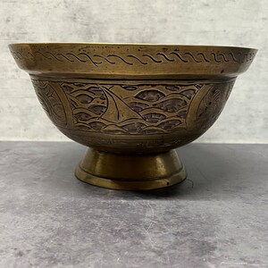 May include: A brass bowl with an intricate design of waves and a boat. The bowl has a wide rim and a pedestal base.