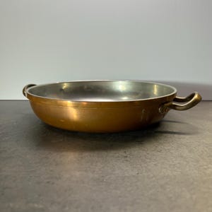 May include: A copper cooking pan with two brass handles. The pan has a shiny, metallic finish and is slightly dented.