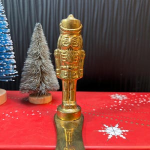 May include: A gold-colored metal nutcracker figurine with a detailed design. The nutcracker is standing on a gold-colored metal base.