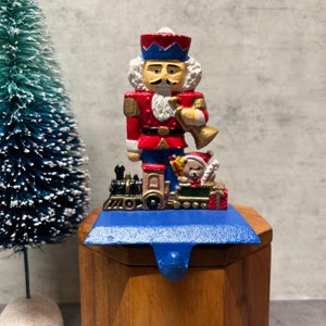 May include: A cast iron stocking holder featuring a painted nutcracker soldier in a red uniform with a blue hat, holding a gold trumpet. At the base, a black steam train engine pulls a green carriage containing a teddy bear and wrapped gifts.