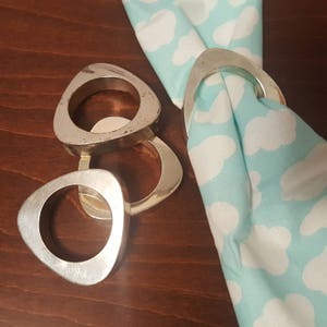 May include: Three silver napkin rings in a triangular shape. The rings are on a wooden surface. One ring is holding a blue and white patterned napkin.