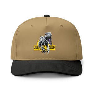 May include: A tan baseball cap with a black brim and a black button on top. The front features a graphic of a grey dinosaur with an open mouth, positioned in front of a yellow banner with the text Jurassic World Dominion printed in black.