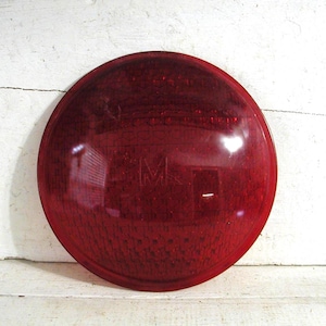 May include: A round, red, vintage automotive tail light lens. The lens is made of a translucent material and has the letters "GMC" embossed on the surface. The lens is designed to fit a specific vehicle model.