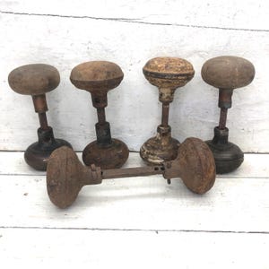 May include: Five antique door knobs with a weathered, rustic appearance. The knobs are various shades of brown and rust, with metal stems and bases. One knob is a handle. The door knobs are arranged on a white wooden surface.