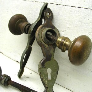 May include: A vintage door hardware set featuring two round brass knobs connected by a threaded metal spindle. The assembly includes an elongated, olive-green painted metal escutcheon plate with a central keyhole and screw holes at the top and bottom.