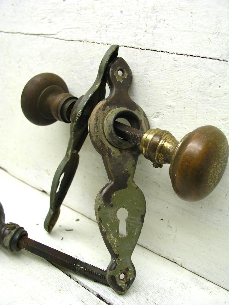 May include: A vintage door hardware set featuring two round brass knobs connected by a threaded metal spindle. The assembly includes an elongated, olive-green painted metal escutcheon plate with a central keyhole and screw holes at the top and bottom.
