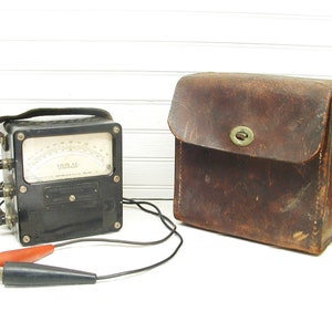 May include: A vintage black analog voltmeter with a leather carrying case. The meter has a dial with the text "VOLTS A.C." and a red and black probe.